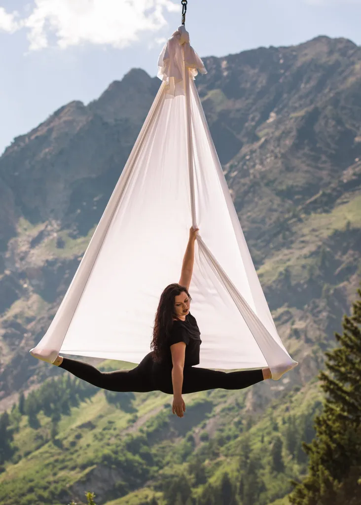 A woman doing aerial yoga in a split pose on a white hammock, suspended against a backdrop of mountains.