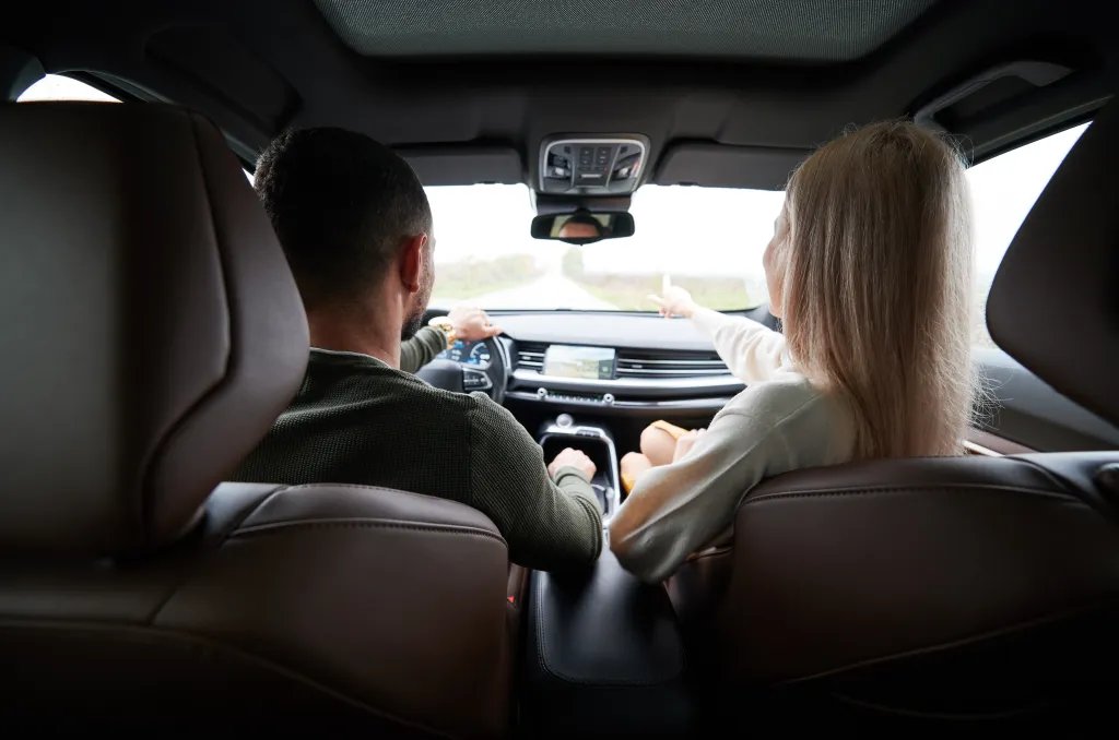 A man driving and a woman pointing ahead from the passenger seat, viewed from the back seats of a car.
