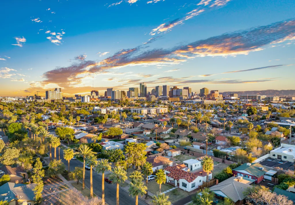 Aerial view of Phoenix, Arizona, downtown skyline with residential areas and palm trees in the foreground, under a sunset sky.