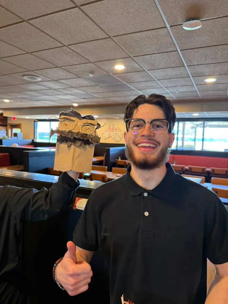 Julian Klein smiles while giving a thumbs-up, next to a puppet made from a paper bag.