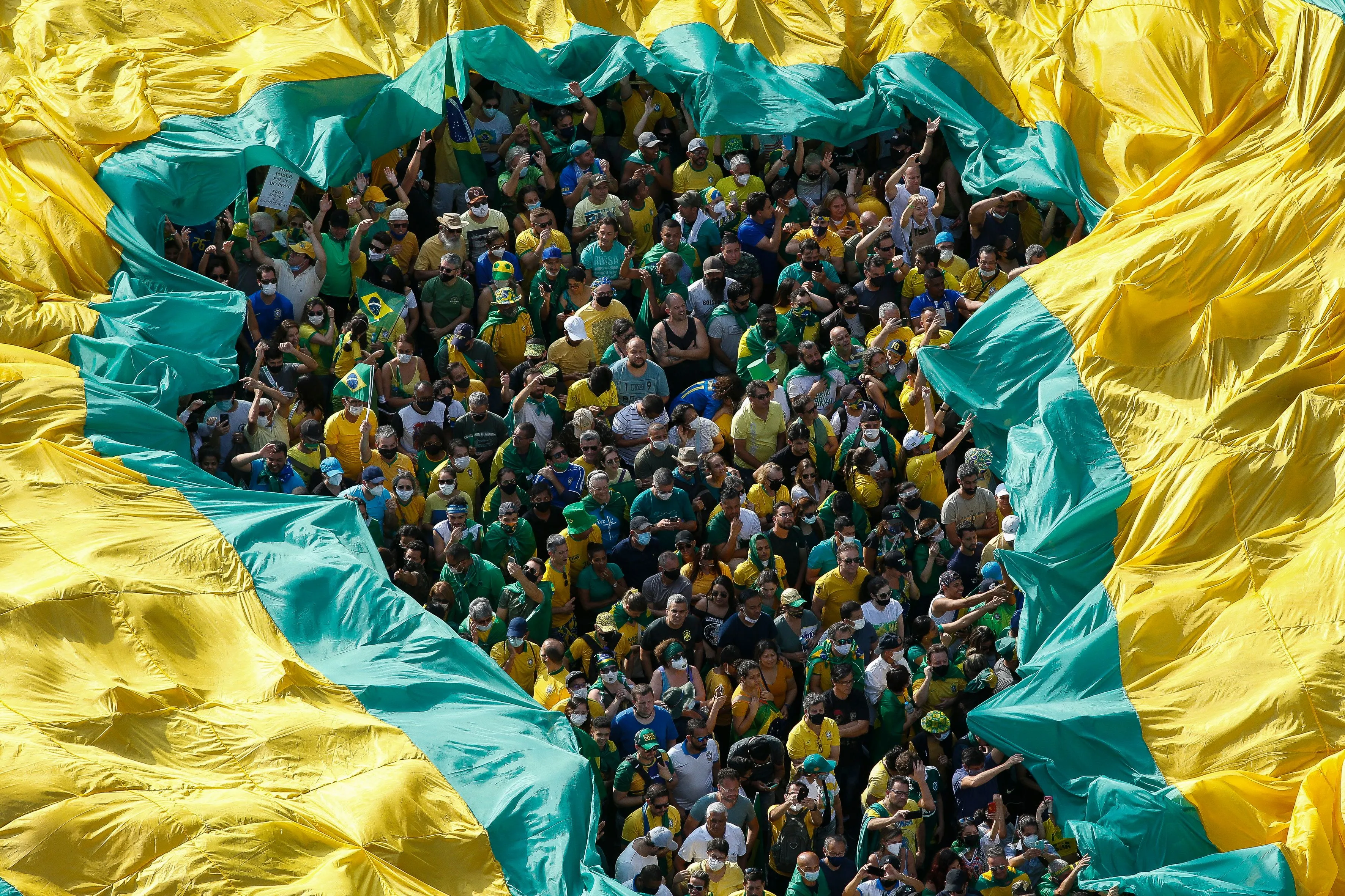 Overhead view of a large crowd dressed in green and yellow gathered beneath a massive piece of fabric in the colors of the Brazilian flag, which is stretched above them with an opening in the center revealing the people below Overhead view of a large crowd dressed in green and yellow gathered beneath a massive piece of fabric in the colors of the Brazilian flag, which is stretched above them with an opening in the center revealing the people below