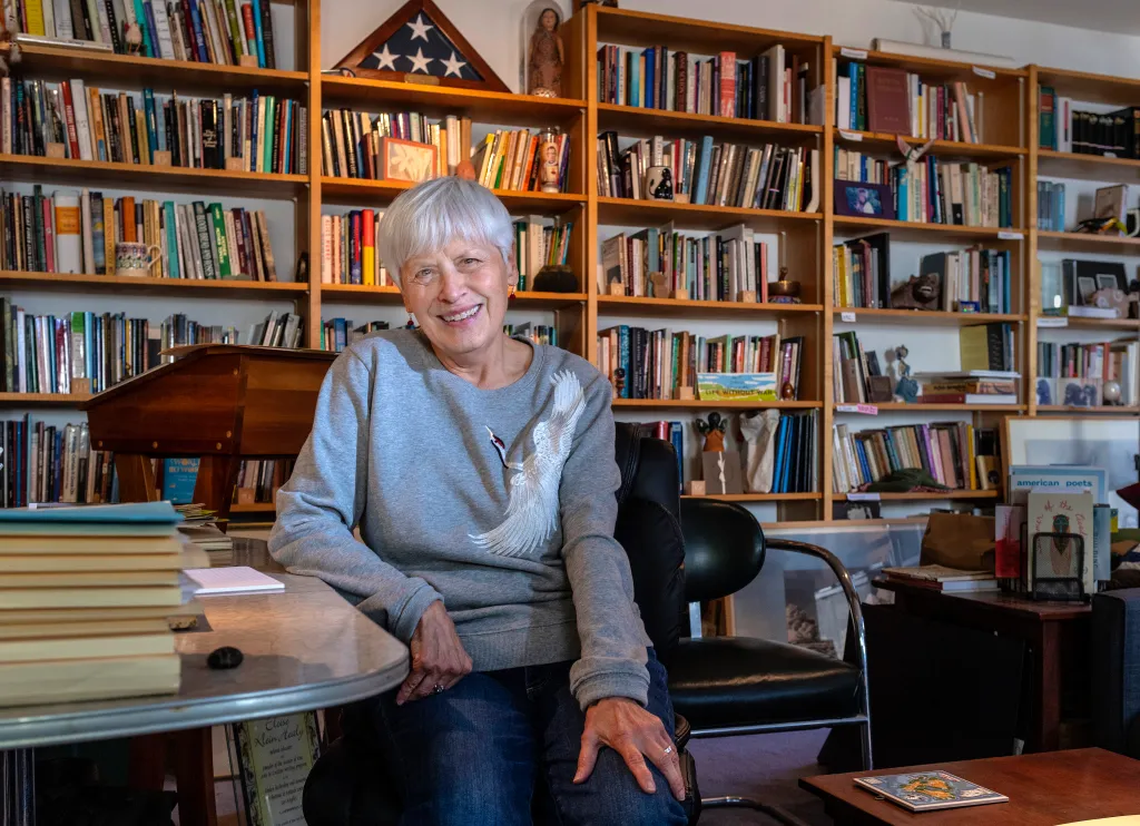 An older woman with short gray hair smiles in front of bookshelves filled with books and a folded American flag.