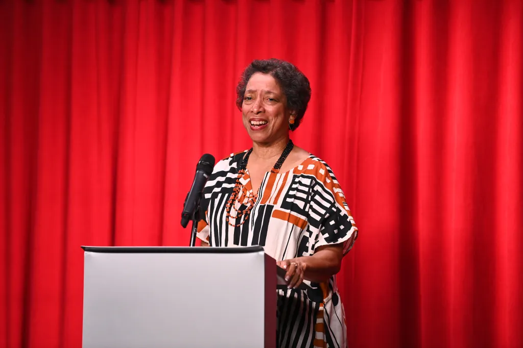 A smiling Black woman in a patterned dress stands at a podium with a microphone, against a red curtain background.