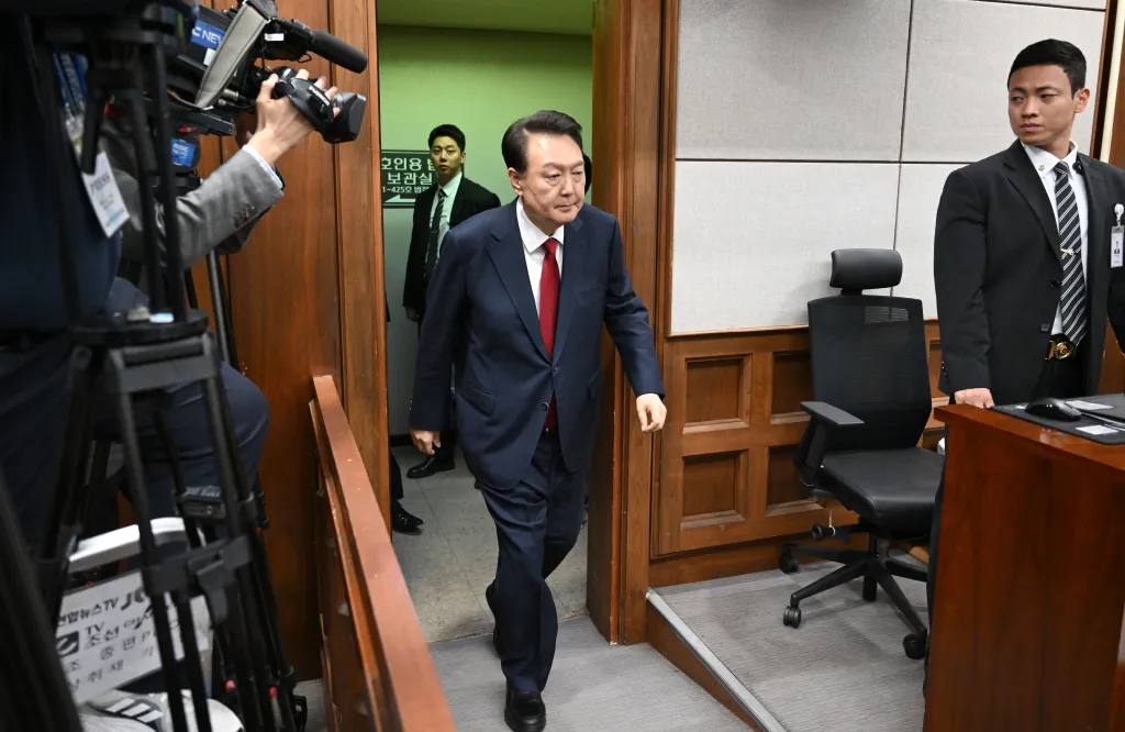 Former President Yoon Suk-yeol walks into the Seoul Central District Court for his criminal trial, with a cameraman and guard nearby.
