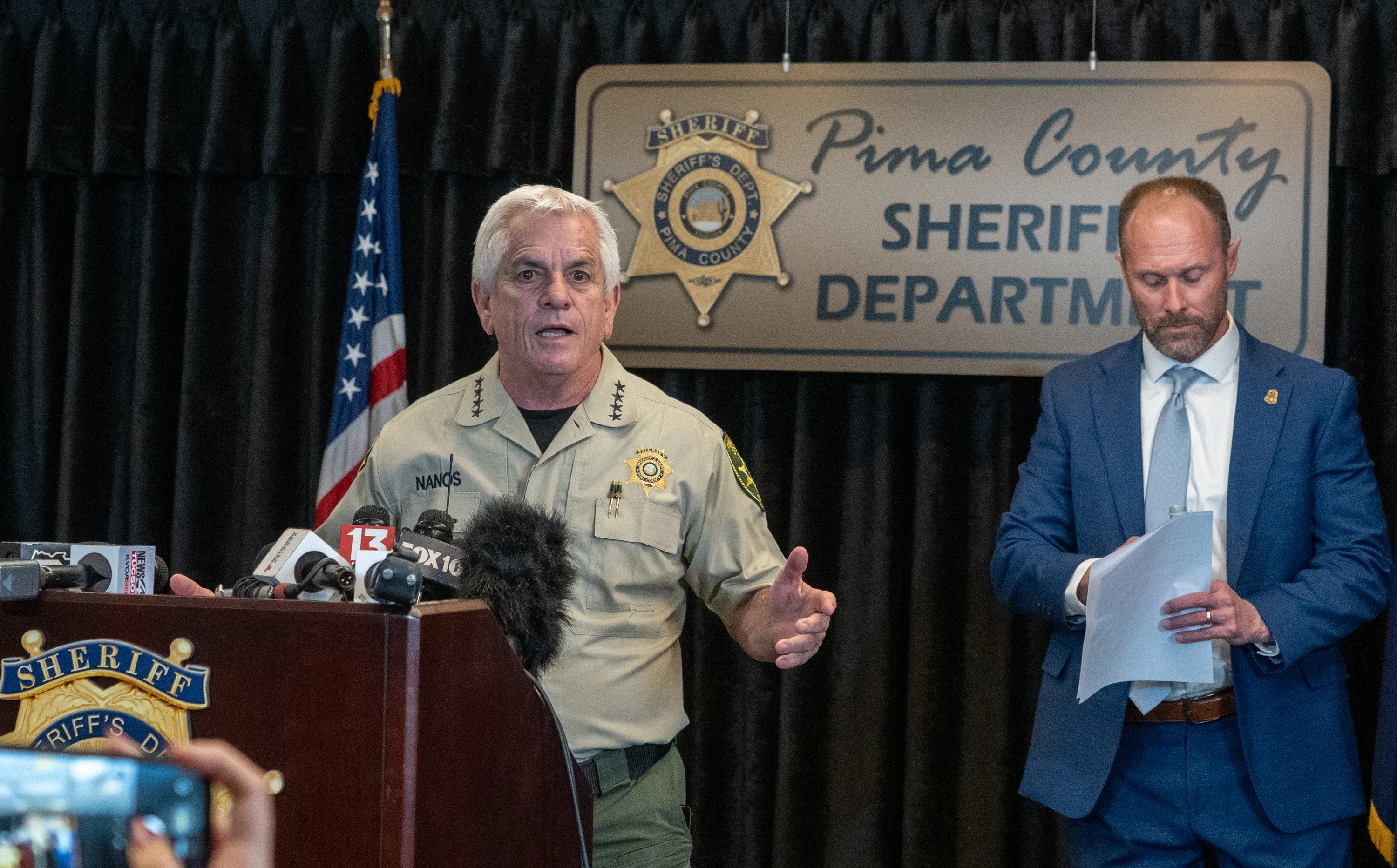 The Pima County sheriff gives a press conference, with a blue-suited FBI agent standing nearby.