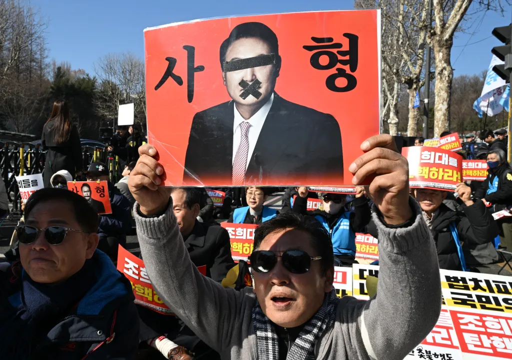 A man holds up a red sign with a picture of a man with tape over his eyes and mouth, with Korean text 