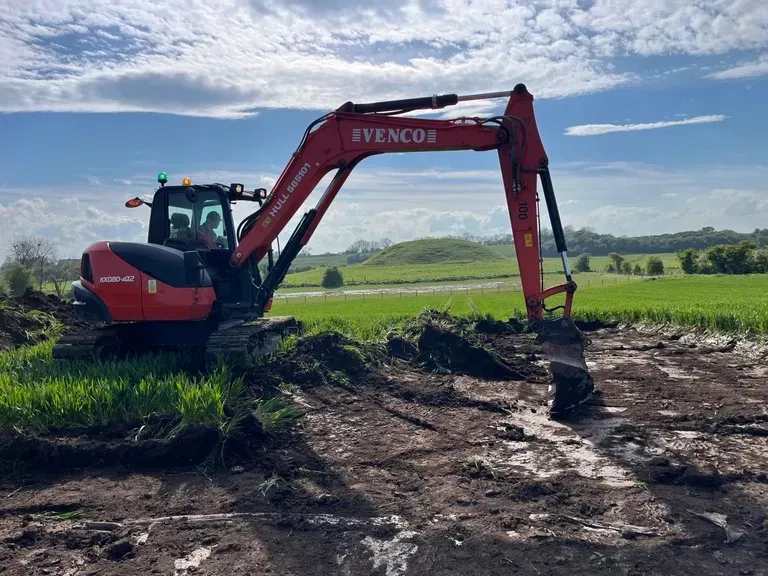 Red and black Venco excavator digging a trench in a grassy field.