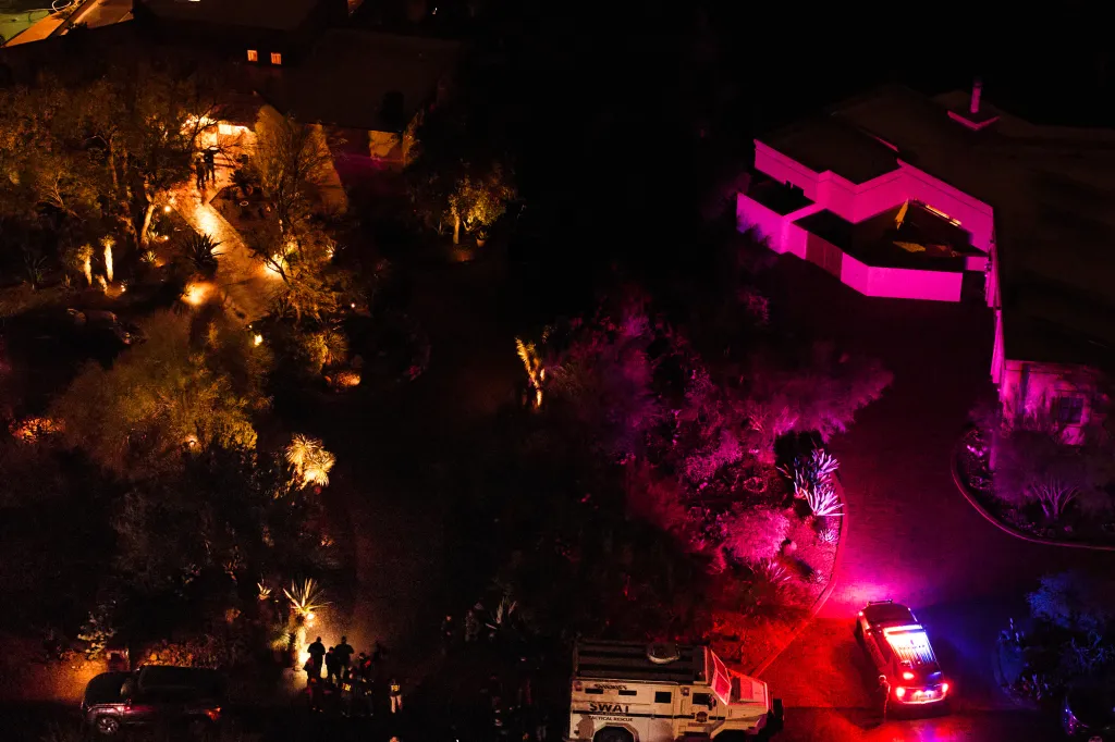 Aerial view of a house and yard at night, with multiple law enforcement vehicles and personnel, illuminated by red, blue, pink, and yellow lights.