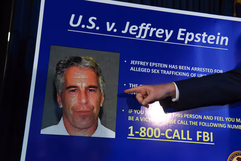 A hand points towards a photo of Jeffrey Epstein on a placard with the heading 'U.S. v. Jeffrey Epstein' and at the bottom it reads '1-800-CALL-FBI'