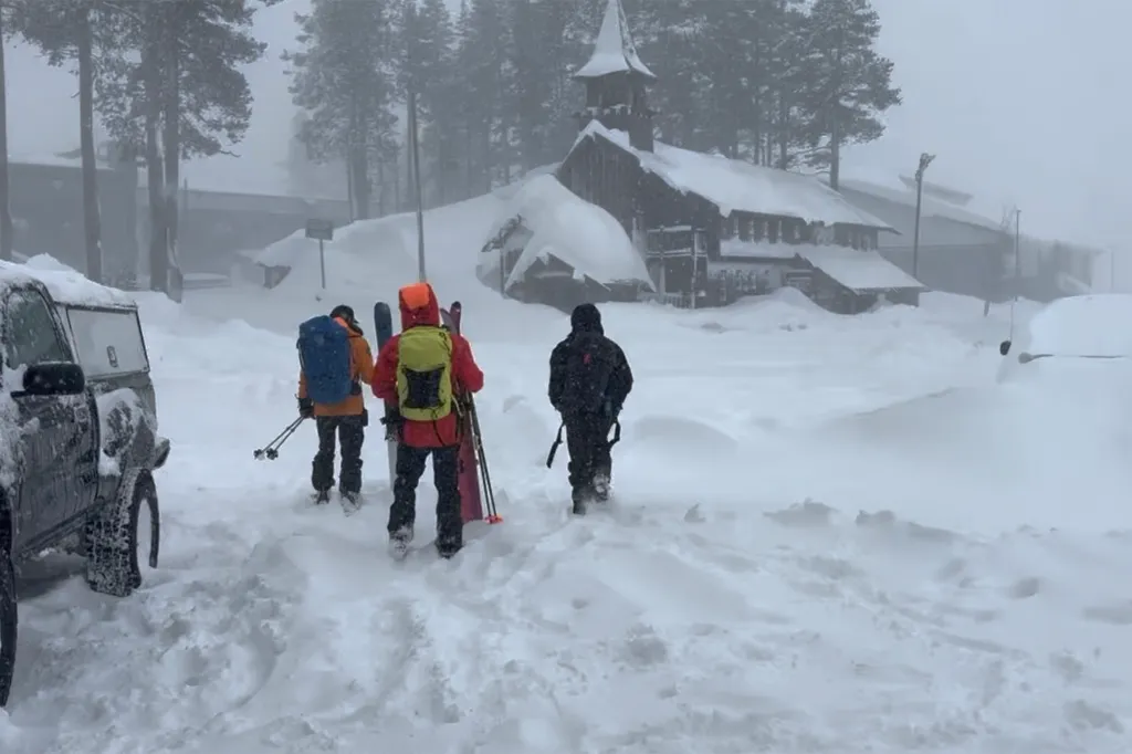 Three people with skis and backpacks walk through deep snow towards a snow-covered building, with a car visible on the left.
