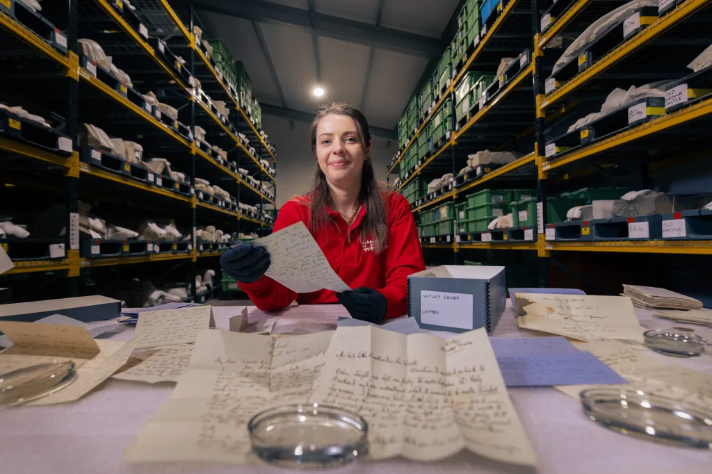 A woman in a red shirt and black gloves holding an old letter, surrounded by other letters on a table, with warehouse shelving full of stored items in the background.