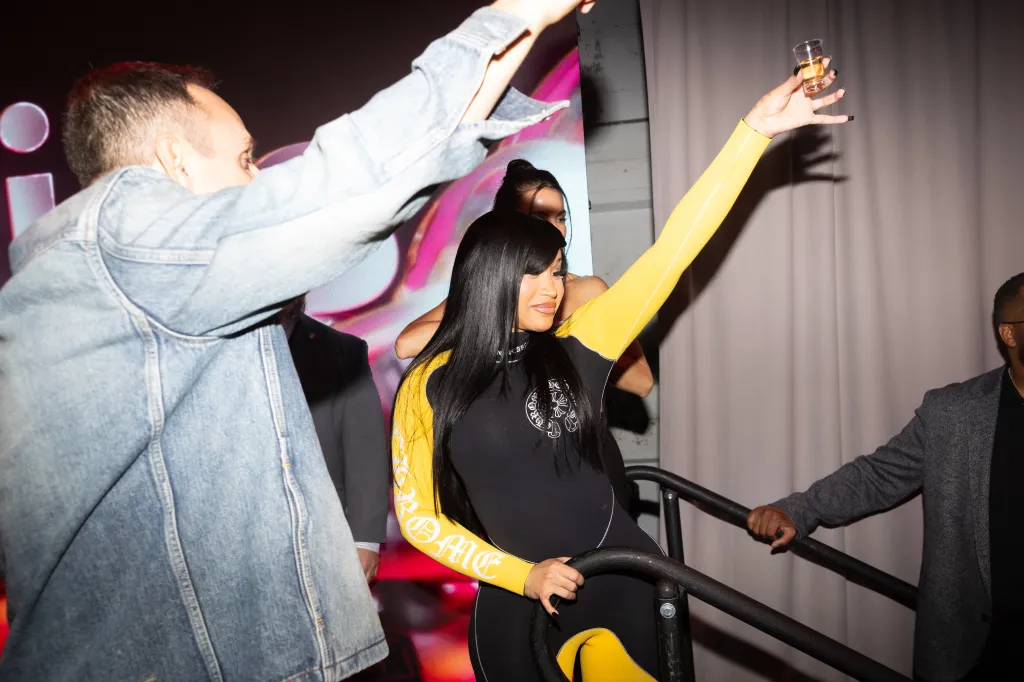 A woman in a black and yellow wetsuit holds a shot glass while another person raises their arms.
