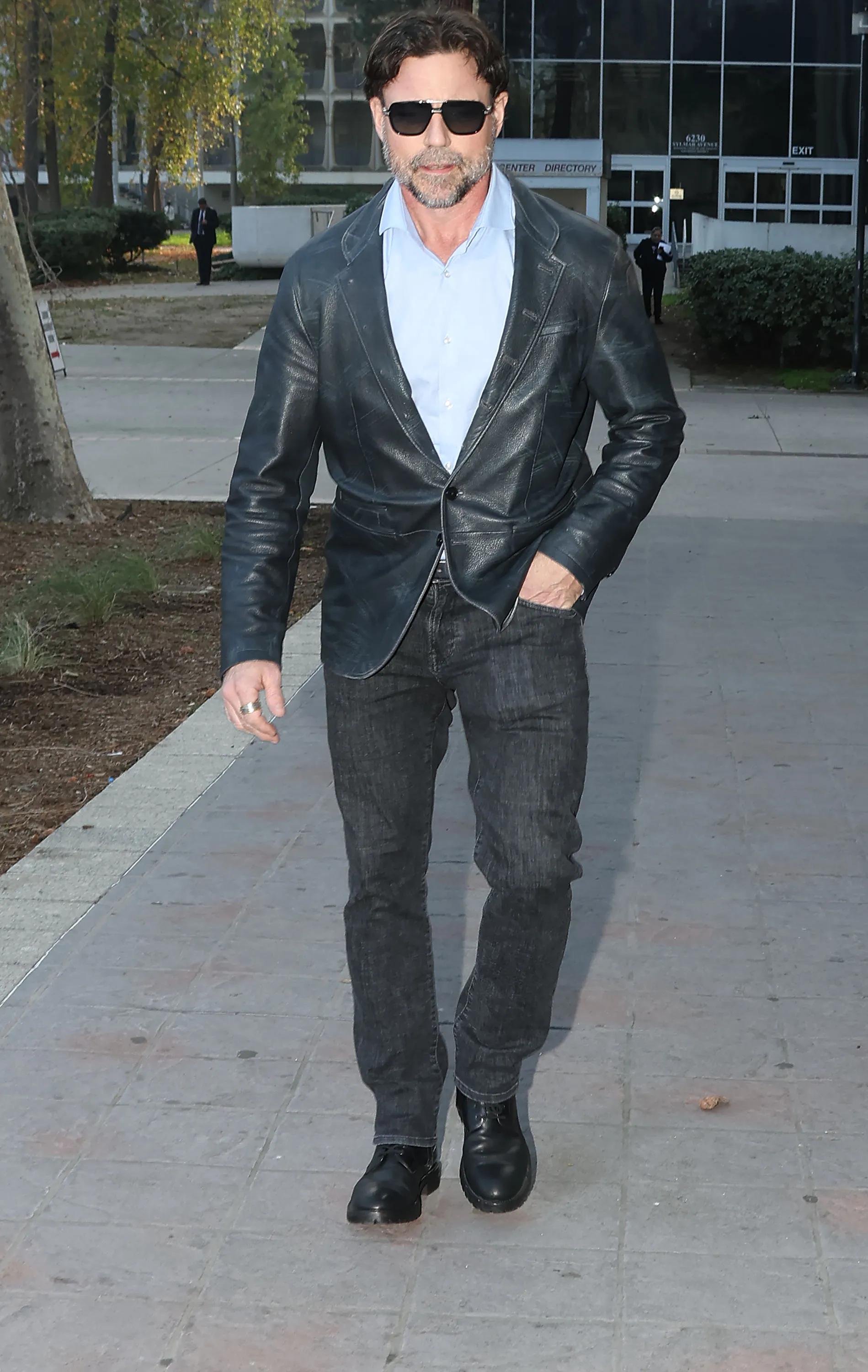 Aaron Phypers in sunglasses, a blue button-up, and a dark leather jacket, walking on a sidewalk.