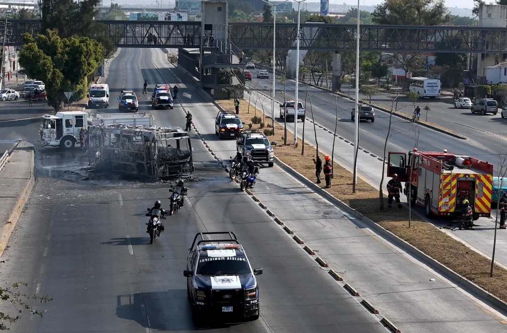 Aerial view of a burnt bus on a highway in Zapopan, Mexico, surrounded by fire trucks, police vehicles, and firefighters.