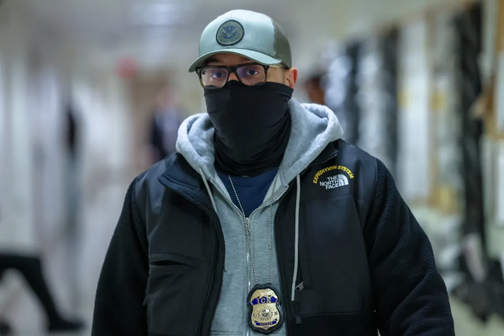 An ICE agent with a mask and hat patrols the halls of immigration court.