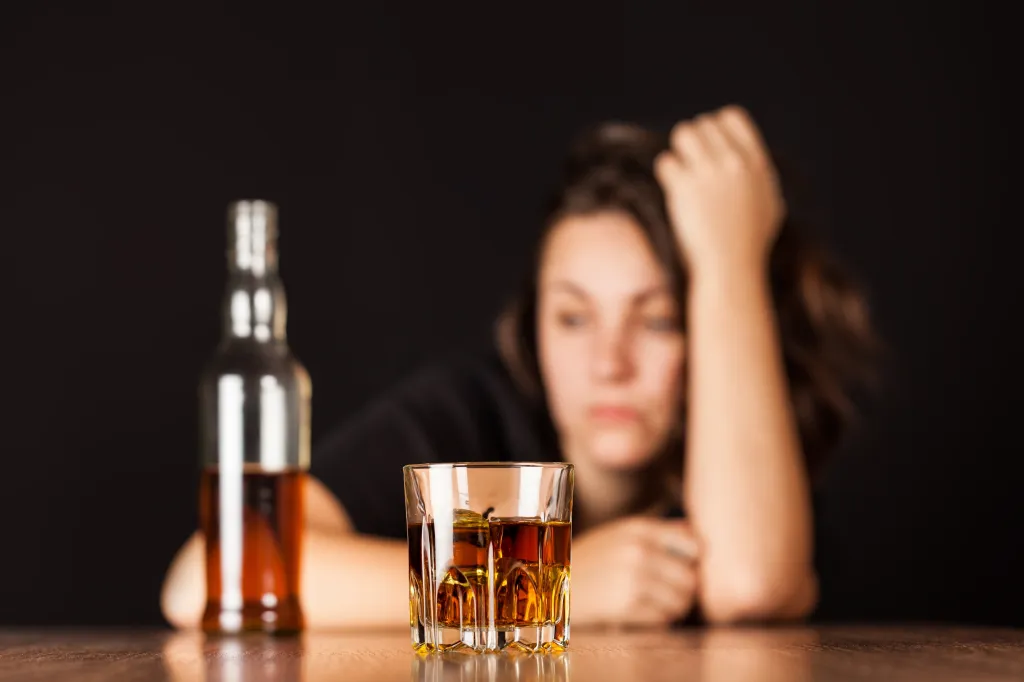A blurred image of a stressed woman with a bottle of alcohol and a glass of whiskey in the foreground.