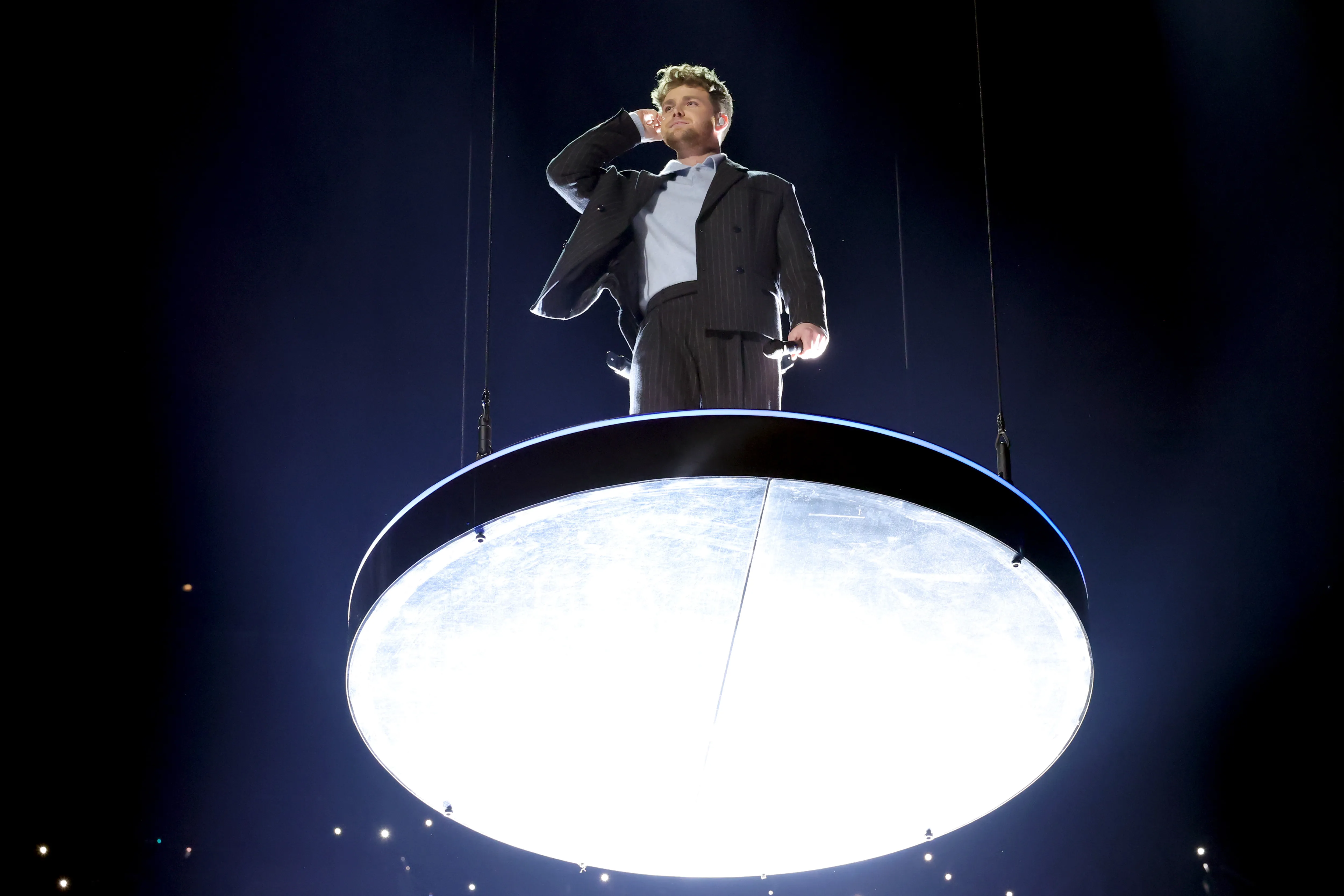 Alex Warren on a circular stage up in the air checking his earpiece