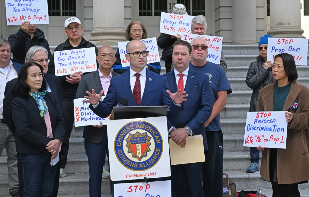 State Assemblyman Edward Ra speaks at a protest against Proposal 1, which aims to reduce ethnic imbalance in specialized high schools, surrounded by protestors holding signs that read 