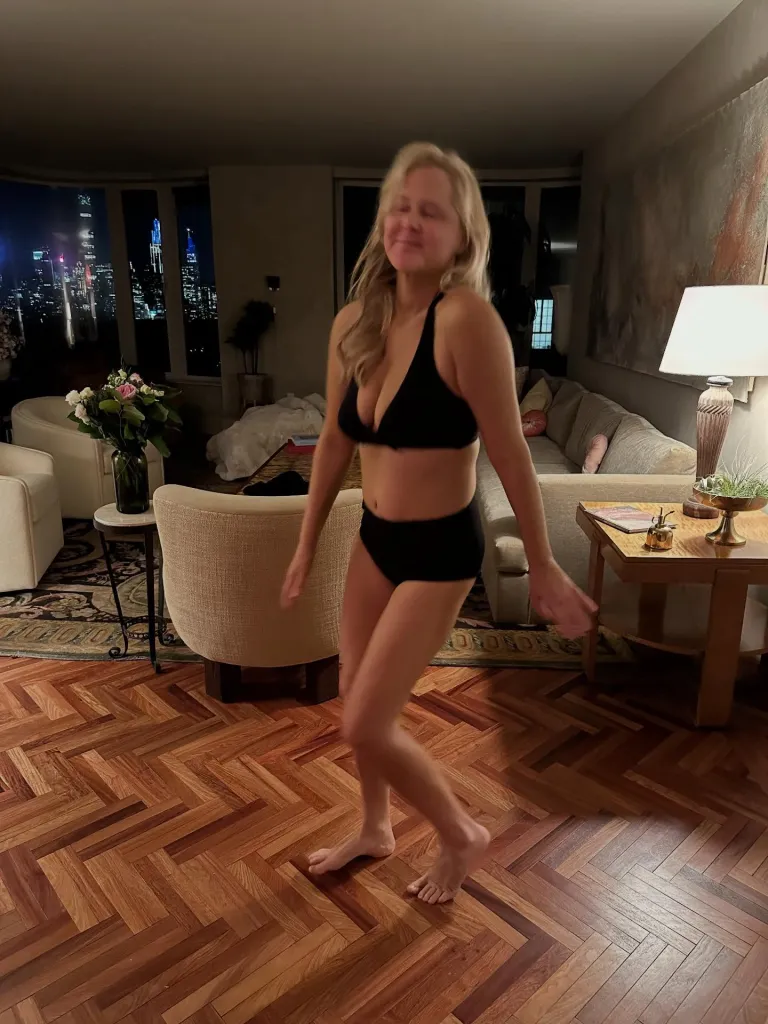Amy Schumer dancing in a black bikini set in a living room with a city night view.