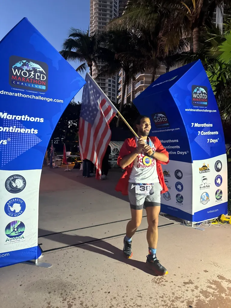 Anthony Simonetti smiling while holding an American flag after completing the World Marathon Challenge, with event banners visible.