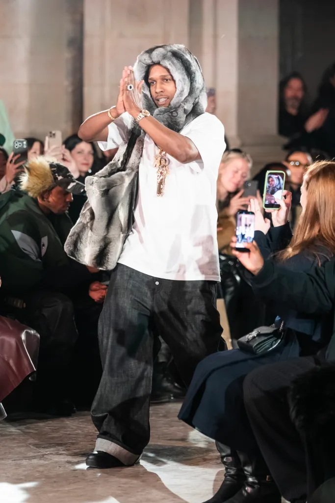 A$AP Rocky walking a runway wearing a white t-shirt, dark jeans, and a fur-lined hood.