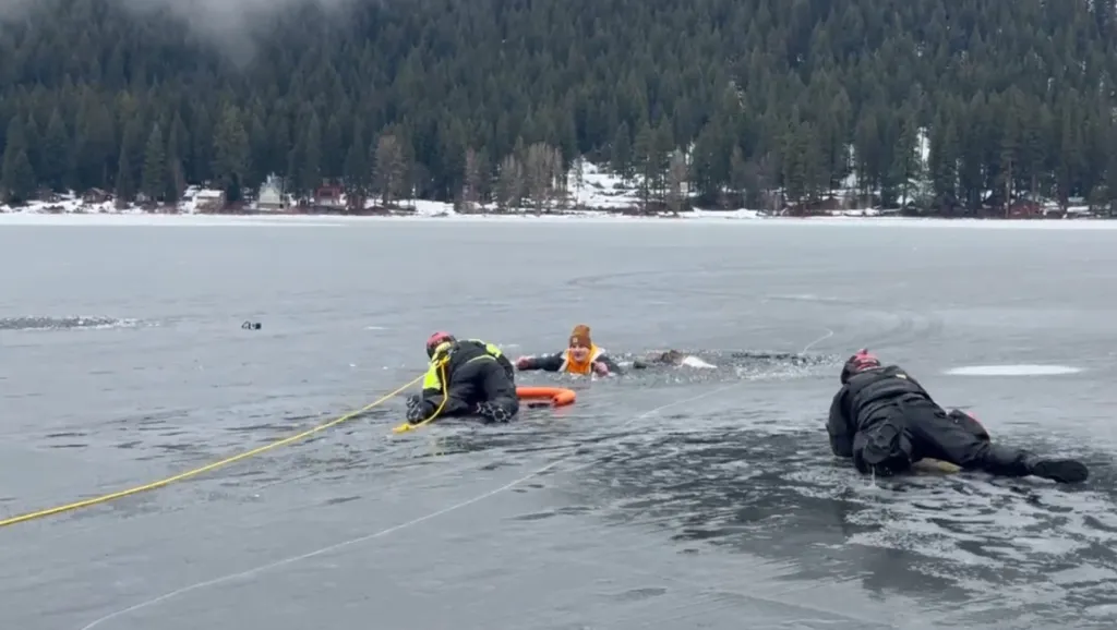 Three people in winter gear performing an ice rescue on a frozen lake with trees and houses in the background.