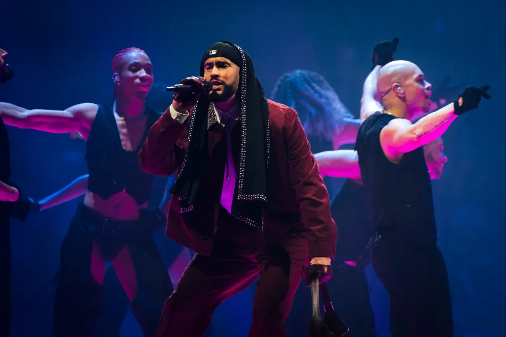Bad Bunny performs at the Barclays Center in a maroon suit and black studded scarf, holding a microphone.