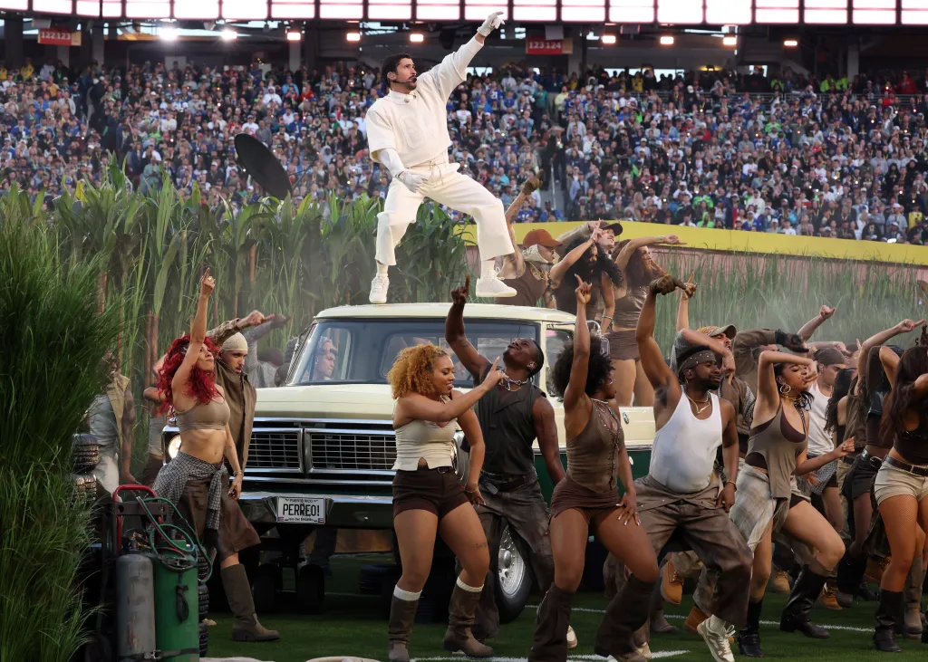 Bad Bunny performing on top of a pickup truck during the Super Bowl Halftime Show, with dancers on stage and a stadium crowd in the background.