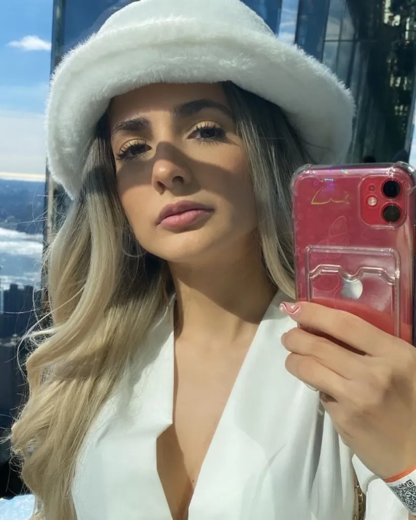 A woman with long blonde hair and a white fluffy hat taking a selfie with a red phone.