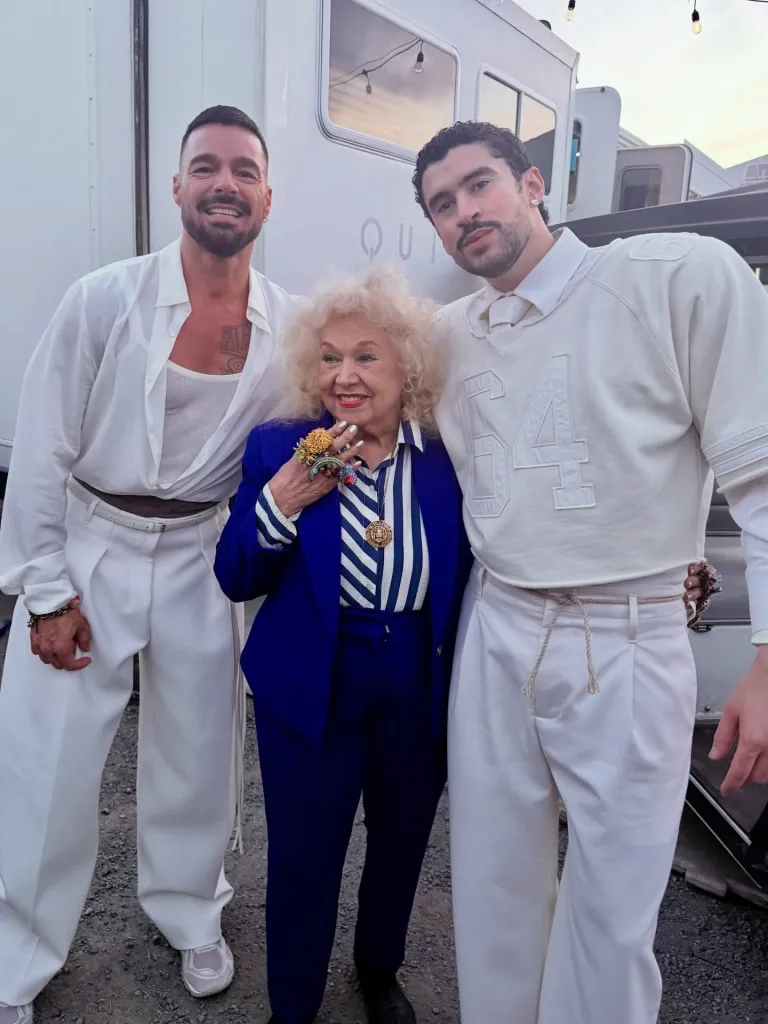 Toñita, a woman with curly blonde hair, wearing a blue pantsuit, stands smiling between two men in white outfits: J Balvin and Bad Bunny.