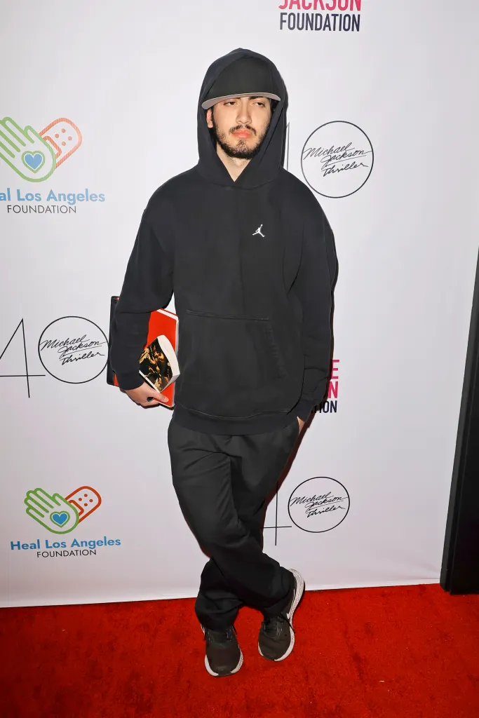 Bigi Jackson poses in a black hoodie and cap on a red carpet.