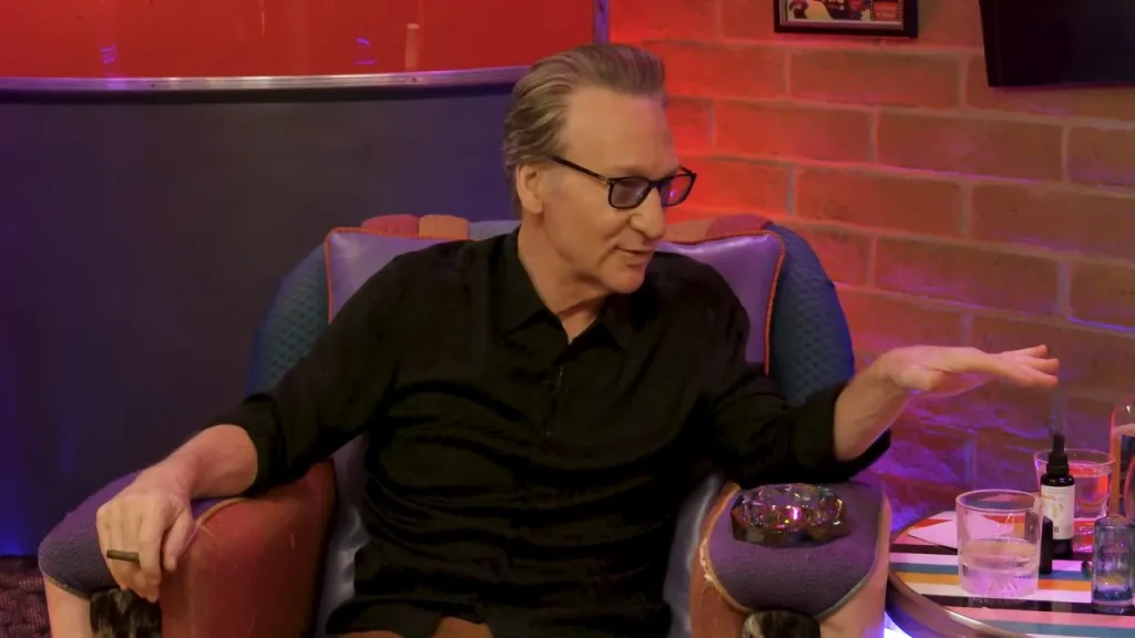 Bill Maher sitting in a chair holding a cigar, with an ashtray and glasses on a table next to him.