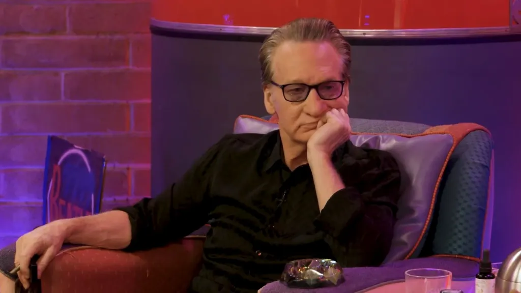 Bill Maher sitting in an armchair, listening intently.