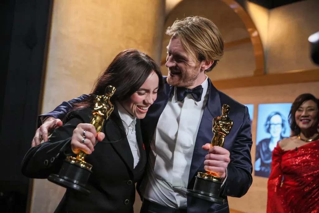 Billie Eilish and Finneas O'Connell backstage at the Academy Awards, each holding an Oscar statue and smiling.