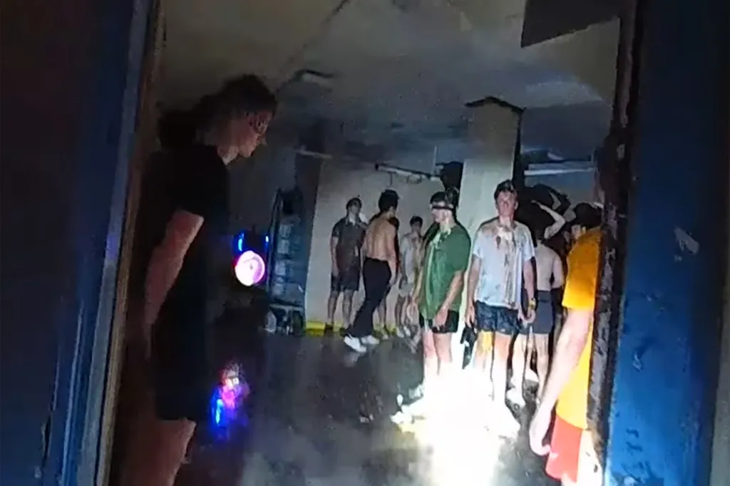 Bodycam footage shows men in a dark, wet basement, many with blindfolds or wearing messy clothes, during an alleged hazing incident.