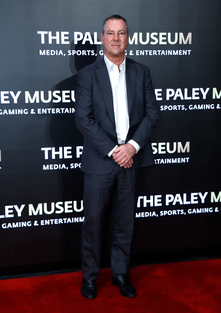 Brad Karp posing for a full-length photo on a red carpet in front of a black backdrop with white text that reads 