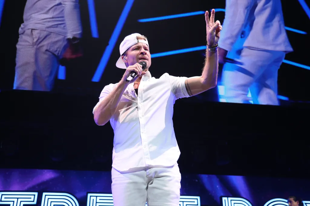 Brian Littrell of the Backstreet Boys performing on stage, wearing a white shirt, shorts, and backwards cap, holding a microphone in one hand and making a peace sign with the other.