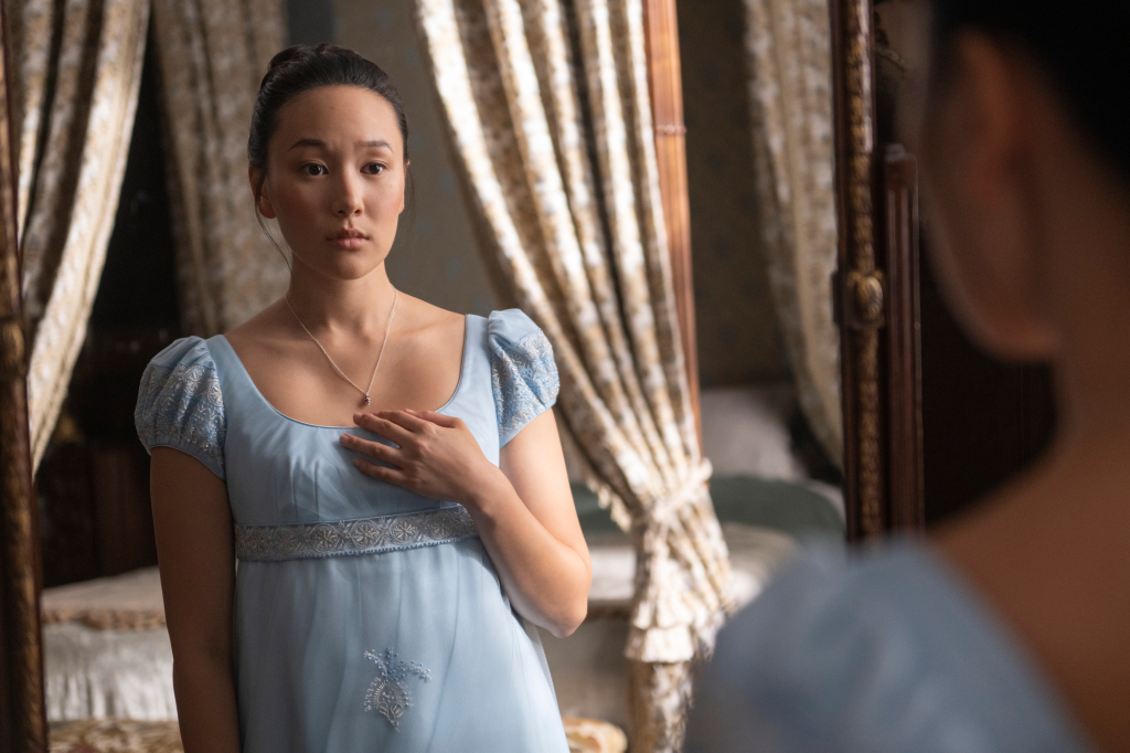Yerin Ha as Sophie Baek in a light blue Regency-era dress looking at her reflection in a mirror.