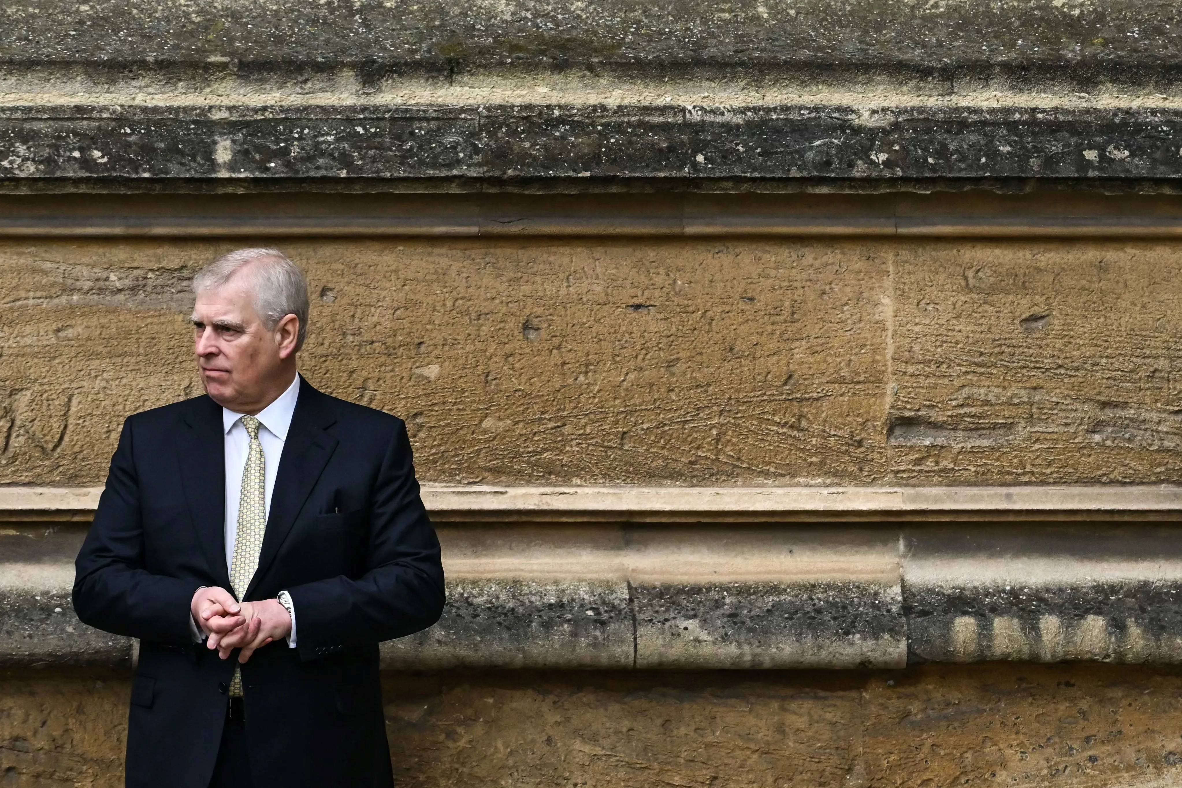 Britain's Prince Andrew, Duke of York, wearing a dark suit and light tie, looks to the left, standing against a stone wall.