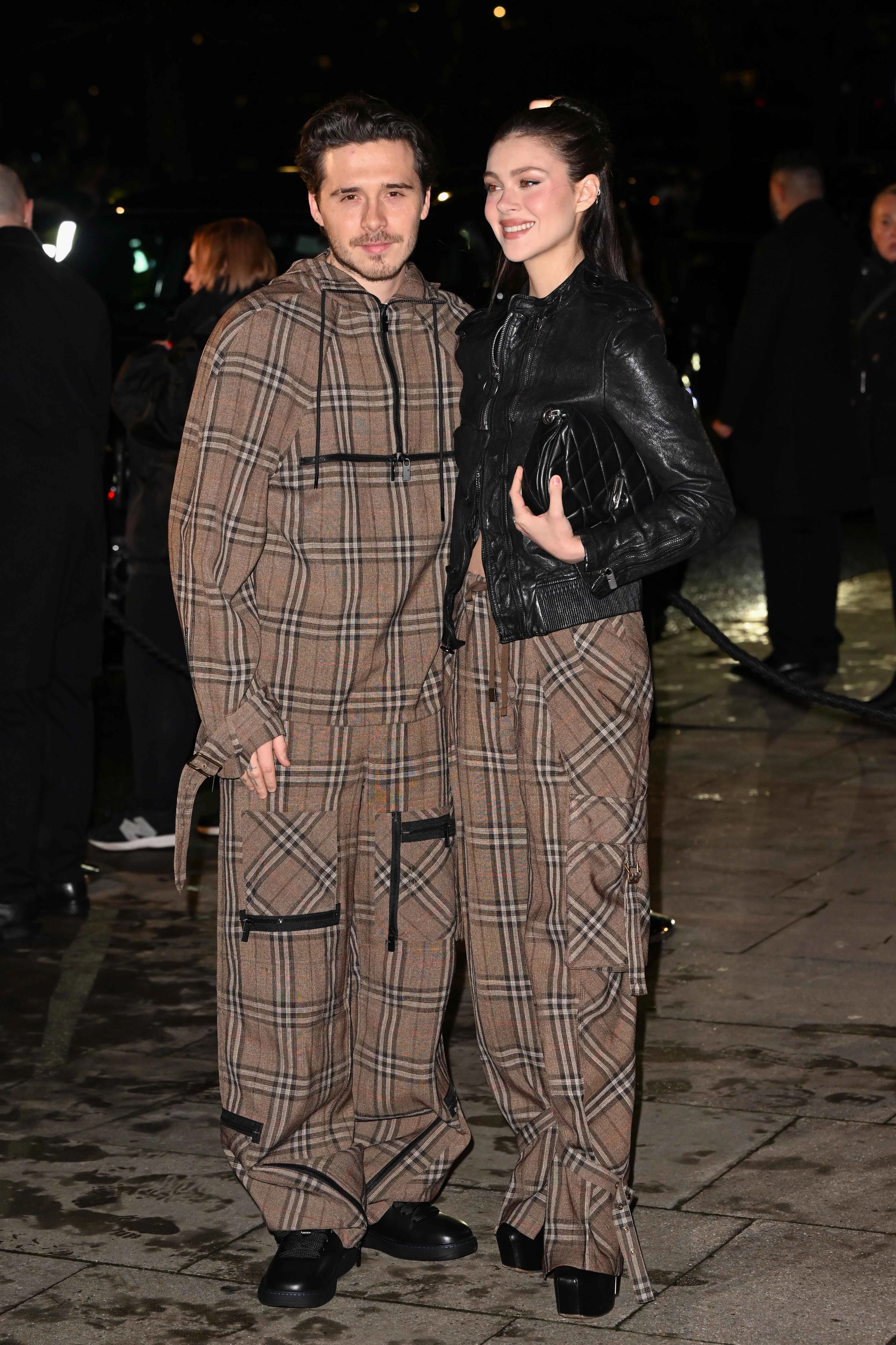 Brooklyn Beckham and Nicola Peltz standing close together, both wearing plaid outfits.