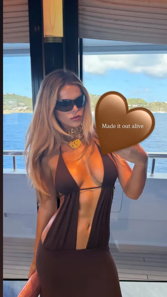 Brooks Nader on a yacht, wearing a brown cut-out dress and sunglasses, with a brown heart emoji covering part of her chest.