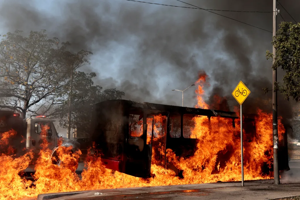 A bus burns in a street with thick black smoke rising into the sky.