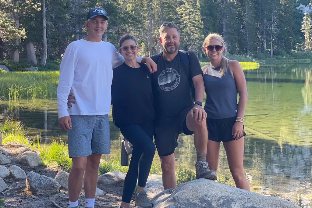 A family of four, two adults and two teenagers, stands next to a lake with pine trees in the background.