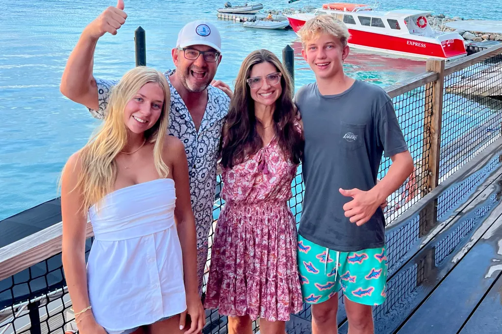 A family of four stands smiling on a dock with boats behind them.