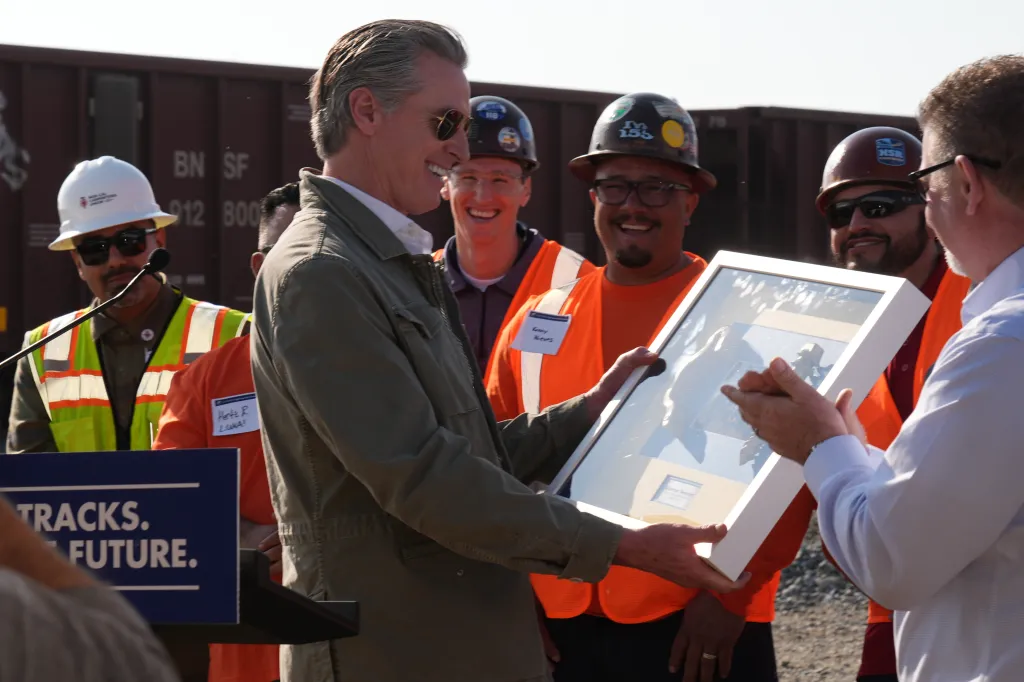 California Gov. Gavin Newsom receives a framed set of golden plated rail fastening clips from a man, as construction workers look on during a news conference about the high-speed rail.