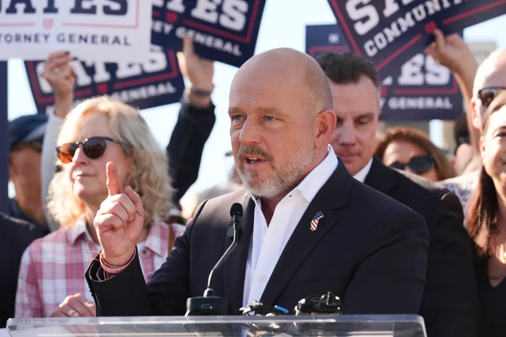 Steve Hilton, a California gubernatorial candidate, speaks at a podium with supporters holding signs for Michael Gates for Attorney General in the background.