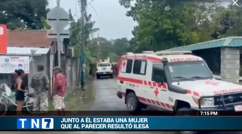 A white ambulance with red markings parked on a street, with people standing in front of shacks and a street sign.