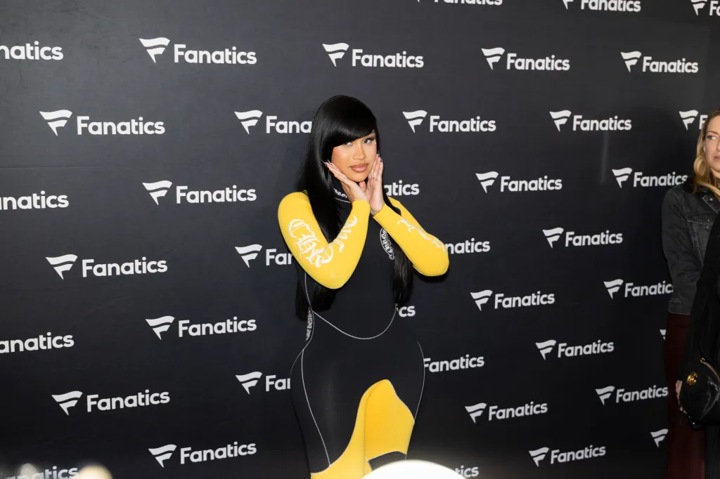 Cardi B posing in a black and yellow outfit with a black backdrop featuring the Fanatics logo.