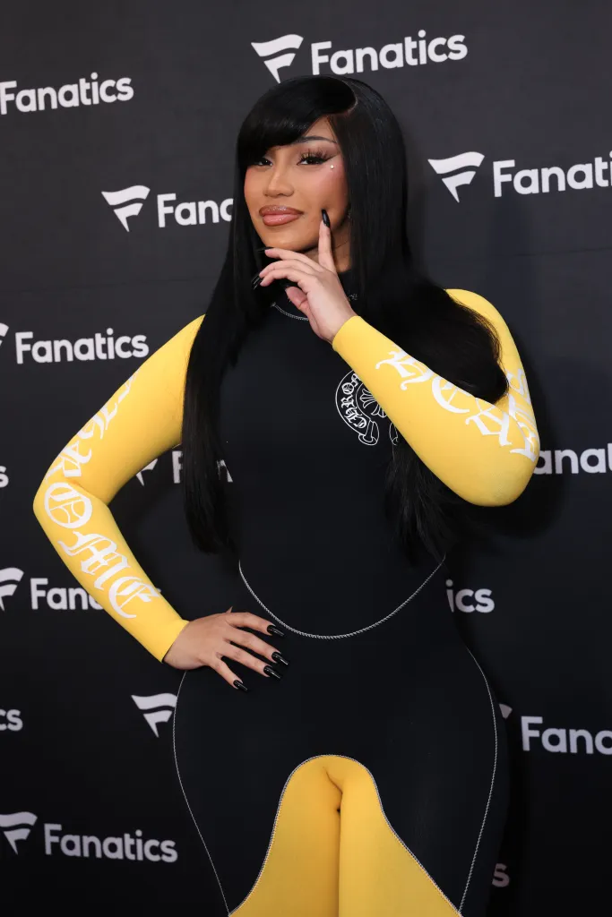 Cardi B posing in a black and yellow outfit with a Fanatics logo backdrop.