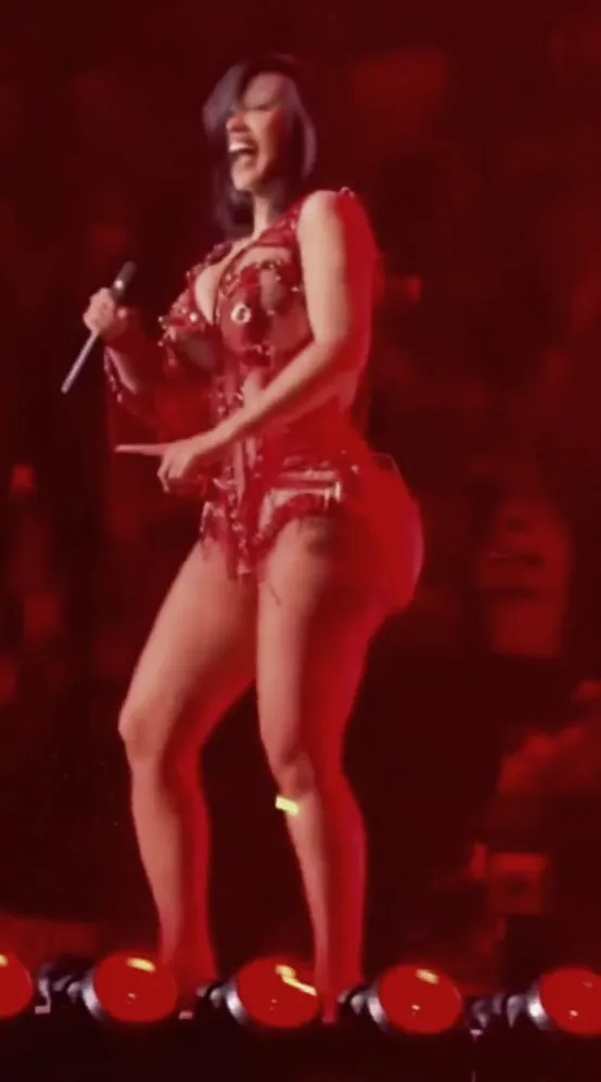 Cardi B performing on stage, dressed in red with a microphone in her left hand, laughing with her mouth wide open.